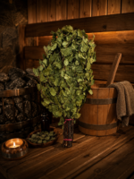 Bērza pirts slota novietota uz koka lāvas tradicionālā pirts interjerā. Blakus redzams koka ķipis, pirts akmeņi, svece un trauks ar kaltētām bērzu lapām.