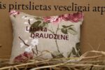 Aromātiskais spilventiņš ar uzrakstu "Labākā draudzene" un ziedu rakstu novietots uz siena fona. Attēlā redzams dekoratīvais spilventiņš ar rozā ziediem un izšūtu uzrakstu.