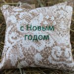 Aromātiskais spilventiņš ar uzrakstu "С Новым годом" novietots uz siena fona.
