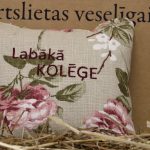 Aromātiskais spilventiņš ar uzrakstu "Labākā kolēģe" un ziedu rakstu novietots uz siena fona. Attēlā redzams dekoratīvais spilventiņš ar rozā ziediem un izšūtu uzrakstu.