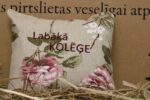 Aromātiskais spilventiņš ar uzrakstu "Labākā kolēģe" un ziedu rakstu novietots uz siena fona. Attēlā redzams dekoratīvais spilventiņš ar rozā ziediem un izšūtu uzrakstu.