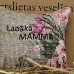 Aromātiskais spilventiņš ar uzrakstu "Labākā mamma" novietots uz siena fona. Attēlā redzams dekoratīvais spilventiņš ar rozā ziedu rakstu un izšūtu uzrakstu, kas izceļ sirsnīgu dāvanas noskaņu.
