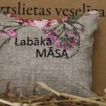Aromātiskais spilventiņš ar uzrakstu "Labākā māsa" novietots uz siena fona. Attēlā redzams dekoratīvais spilventiņš ar izšūtu uzrakstu un maigu ziedu rakstu rozā toņos.