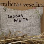 Aromātiskais spilventiņš ar uzrakstu "Labākā meita" novietots uz siena fona. Attēlā redzams dekoratīvs spilventiņš ar izšūtu uzrakstu un maigiem ziedu motīviem, radot siltu un mājīgu noskaņu.