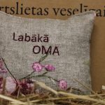 Aromātiskais spilventiņš ar uzrakstu "Labākā oma" novietots uz siena fona. Attēlā redzams dekoratīvais spilventiņš ar izšūtu uzrakstu un maigu ziedu rakstu rozā toņos.