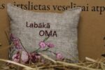 Aromātiskais spilventiņš ar uzrakstu "Labākā oma" novietots uz siena fona. Attēlā redzams dekoratīvais spilventiņš ar izšūtu uzrakstu un maigu ziedu rakstu rozā toņos.