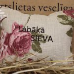 Aromātiskais spilventiņš ar uzrakstu "Labākā sieva" novietots uz siena fona. Attēlā redzams dekoratīvs spilventiņš ar izšūtu uzrakstu un lieliem rozā ziedu motīviem, radot siltu un romantisku noskaņu.