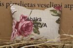 Aromātiskais spilventiņš ar uzrakstu "Labākā sieva" novietots uz siena fona. Attēlā redzams dekoratīvs spilventiņš ar izšūtu uzrakstu un lieliem rozā ziedu motīviem, radot siltu un romantisku noskaņu.