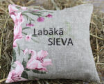 Dekoratīvs lina spilventiņš ar izšuvumu "Labākā SIEVA" un ziedu rakstu novietots uz siena fona. Spilventiņam ir cilpiņa pakāršanai un dabīga lina auduma faktūra.