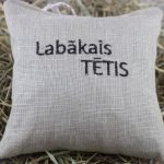 Dekoratīvs lina spilventiņš ar izšuvumu "Labākais TĒTIS" novietots uz siena fona. Spilventiņam ir cilpiņa pakāršanai un dabīga lina auduma faktūra.
