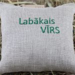 Dekoratīvs lina spilventiņš ar izšuvumu "Labākais VĪRS" novietots uz siena fona. Spilventiņam ir cilpiņa pakāršanai un dabīga lina auduma faktūra.
