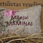 Aromātiskais spilventiņš ar uzrakstu "Mīļajai māmiņai" novietots uz siena fona. Attēlā redzams dekoratīvs spilventiņš ar izšūtu uzrakstu un maigu ziedu rakstu rozā toņos, radot siltu un mājīgu noskaņu.