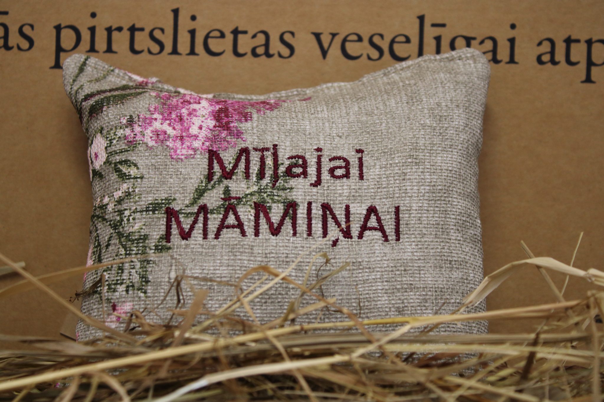 Aromātiskais spilventiņš ar uzrakstu "Mīļajai māmiņai" novietots uz siena fona. Attēlā redzams dekoratīvs spilventiņš ar izšūtu uzrakstu un maigu ziedu rakstu rozā toņos, radot siltu un mājīgu noskaņu.