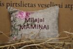 Aromātiskais spilventiņš ar uzrakstu "Mīļajai māmiņai" novietots uz siena fona. Attēlā redzams dekoratīvs spilventiņš ar izšūtu uzrakstu un maigu ziedu rakstu rozā toņos, radot siltu un mājīgu noskaņu.