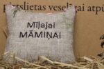Aromātiskais spilventiņš ar uzrakstu "Mīļajai māmiņai" novietots uz siena fona. Attēlā redzams dekoratīvais spilventiņš ar izšūtu uzrakstu un maigu ziedu rakstu.