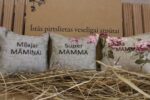 Trīs aromātiskie spilventiņi ar uzrakstiem "Mīļajai māmiņai", "Super mamma" un "Labākā mamma" novietoti uz siena fona. Attēlā redzami dekoratīvi spilventiņi ar ziedu rakstiem, kas veido sirsnīgu dāvanu kompozīciju mammai.