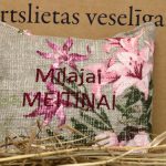 Aromātiskais spilventiņš ar uzrakstu "Mīļajai meitiņai" un rozā ziedu rakstu novietots uz siena fona. Attēlā redzams dekoratīvais spilventiņš ar izšūtu uzrakstu un košu ziedu motīvu.