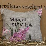 Aromātiskais spilventiņš ar uzrakstu "Mīļajai sieviņai" un ziedu rakstu novietots uz siena fona. Attēlā redzams dekoratīvais spilventiņš ar izšūtu uzrakstu un ceriņu ziedu motīvu.