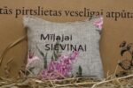 Aromātiskais spilventiņš ar uzrakstu "Mīļajai sieviņai" un ziedu rakstu novietots uz siena fona. Attēlā redzams dekoratīvais spilventiņš ar izšūtu uzrakstu un ceriņu ziedu motīvu.
