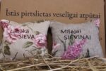 Divi aromātiskie spilventiņi ar uzrakstiem "Labākā sieva" un "Mīļajai sieviņai" novietoti uz siena fona. Attēlā redzami dekoratīvi spilventiņi ar ziedu rakstiem un izšūtiem uzrakstiem.