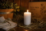 Dekoratīva balta svece novietota uz koka lāvas pirts interjerā. Fonā redzams koka ķipis, pirts slota, linu dvielis un kaste ar uzrakstu "Īstas pirts lietas".