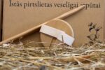 Apaļas pirts birstes aizmugure ar regulējamu auduma siksniņu un garu koka kātu novietota uz siena fona. Attēlā redzama gluda koka virsma un ergonomiska konstrukcija ērtai lietošanai.