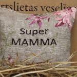 Aromātiskais spilventiņš ar uzrakstu "Super mamma" novietots uz siena fona. Attēlā redzams dekoratīvs spilventiņš ar izšūtu uzrakstu un maigu rozā ziedu rakstu, radot siltu un mājīgu noskaņu.