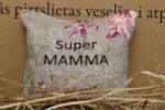 Aromātiskais spilventiņš ar uzrakstu "Super mamma" novietots uz siena fona. Attēlā redzams dekoratīvs spilventiņš ar izšūtu uzrakstu un maigu rozā ziedu rakstu, radot siltu un mājīgu noskaņu.