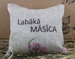 Aromātiskais spilventiņš ar uzrakstu "Labākā MĀSĪCA" novietots uz siena fona.