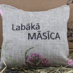 Aromātiskais spilventiņš ar uzrakstu "Labākā MĀSĪCA" novietots uz siena fona.
