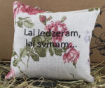 Aromātiskais spilventiņš ar uzrakstu "Lai iedzeram, lai svinam." novietots uz siena fona.