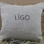 Aromātiskais spilventiņš ar uzrakstu "LĪGO" novietots uz siena fona.