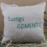 Aromātiskais spilventiņš ar uzrakstu "Lustīgu DZIMENĪTI!" novietots uz siena fona.