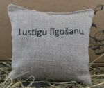 Aromātiskais spilventiņš ar uzrakstu "Lustīgu līgošanu" novietots uz siena fona.