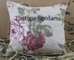 Aromātiskais spilventiņš ar uzrakstu "Lustīgu līgošanu!" novietots uz siena fona.