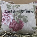 Aromātiskais spilventiņš ar uzrakstu "Lustīgu līgošanu!" novietots uz siena fona.