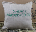 Aromātiskais spilventiņš ar uzrakstu "Sveiciens VĀRDINSVĒTKOS!" novietots uz siena fona.
