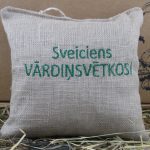 Aromātiskais spilventiņš ar uzrakstu "Sveiciens VĀRDINSVĒTKOS!" novietots uz siena fona.