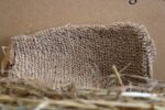 Dabīga lufa pirts dūrainis novietots uz siena fona. Attēlā redzama dūraiņa atvērtā puse, izceļot tā apjomu, formu un dabīgās šķiedras tekstūru.
