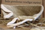 Dabīga lufa pirts dūrainis novietots uz siena fona. Attēlā redzama dūraiņa iekšpuse un plašā atvēruma forma, izceļot dabīgo šķiedru pinumu un auduma cilpas.