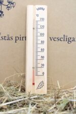 Pirts termometrs uz salmu fona. Attēlā redzams koka termometrs ar temperatūras skalu līdz 140 °C un dekoratīvu lapas motīvu apakšdaļā.