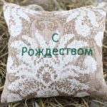 Aromātiskais spilventiņš ar uzrakstu "С Рождеством" novietots uz siena fona.
