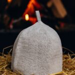 Pelēka lina pirts cepure kā galvenais akcents pirts telpā, novietota uz koka virsmas, fonā silta uguns gaisma