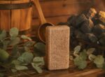 Ciedra aromāta sāls bloks novietots siltā pirts atmosfērā uz koka lāvas. Attēlā redzams brūngans sāls bloks kopā ar eikalipta lapām, koka ķipīti, kausiņu un krāsns akmeņiem fonā.