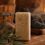 Eikalipta aromāta sāls bloks novietots siltā pirts atmosfērā uz koka lāvas. Attēlā redzams gaišs sāls bloks kopā ar eikalipta lapām, lina audumu, koka ķipīti un krāsns akmeņiem fonā.