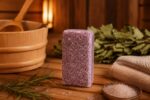 Rozmarīna aromāta sāls bloks novietots siltā pirts atmosfērā uz koka lāvas. Attēlā redzams violetīgs sāls bloks kopā ar rozmarīna zariem, jūras sāls kristāliem, sarullētu dvieli un bērza zariem fonā.