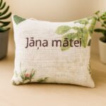 Aromātiskais auduma spilventiņš ar uzrakstu “Jāņa mātei”, novietots uz gaišas koka virsmas