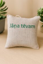Aromātiskais auduma spilventiņš ar uzrakstu “Jāņa tēvam”, novietots uz gaišas koka virsmas