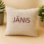 Aromātiskais auduma spilventiņš ar uzrakstu “JĀNIS”, novietots uz gaišas koka virsmas