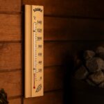 Koka pirts termometrs ar temperatūras skalu piestiprināts pie koka sienas blakus pirts krāsns akmeņiem